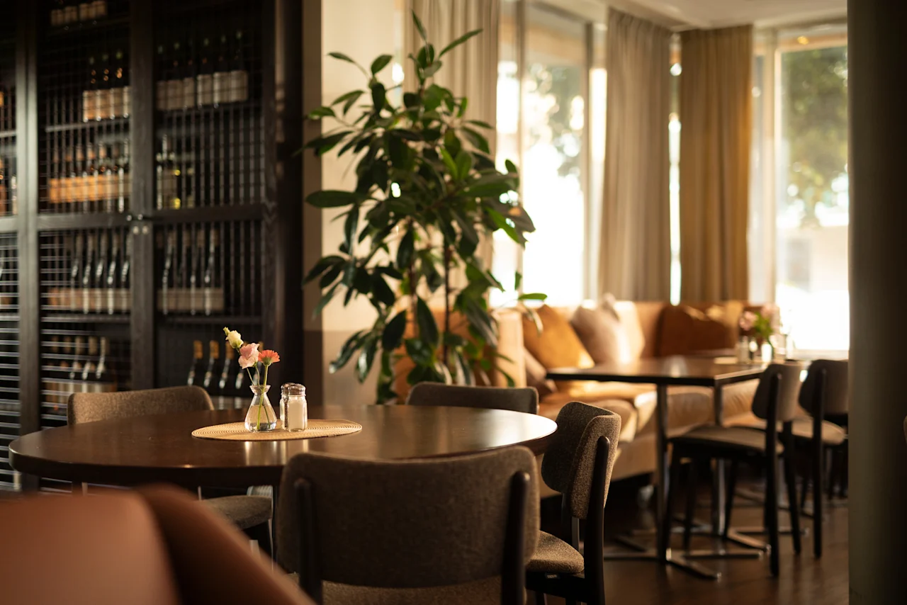 En varm och inbjudande restaurang eller lounge med dukade bord, en stor växt och en vinförvaring, badad i naturligt ljus.