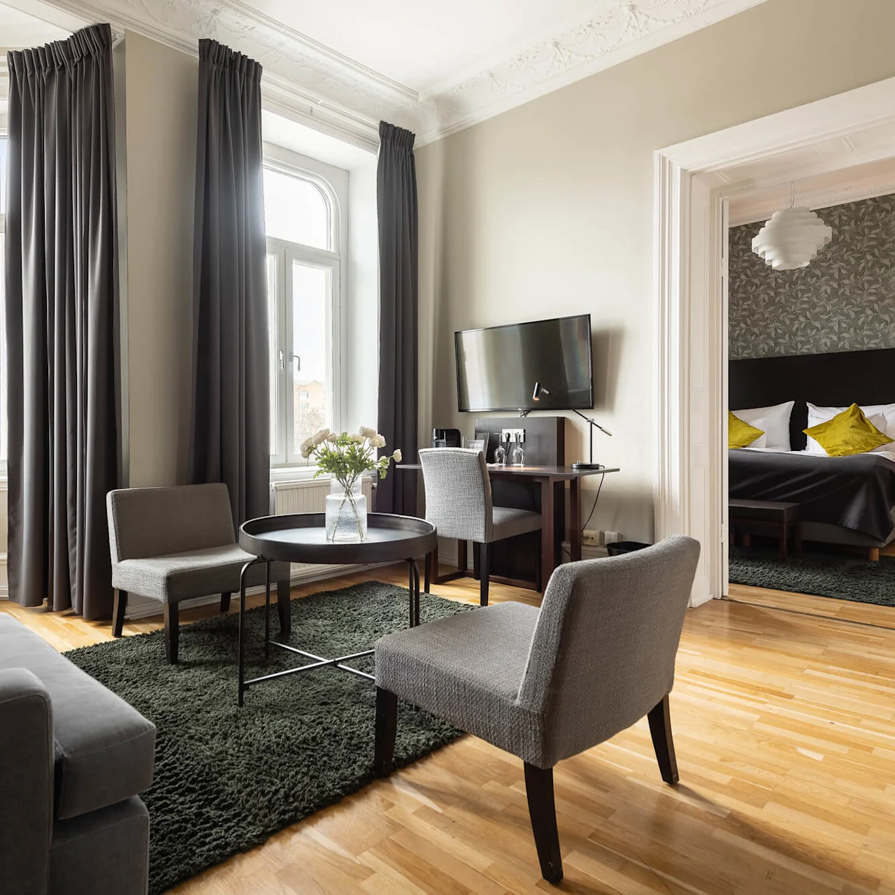 A modern living room with a sofa, chairs, and a TV, transitioning to a bedroom in the background, lit by daylight from large windows.