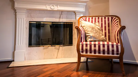 A striped armchair with a shiny pillow sits beside a decorative white marble fireplace in a warmly lit room with wooden flooring.