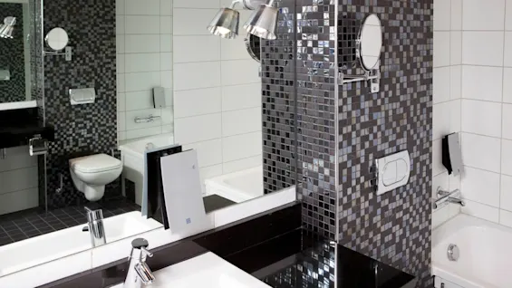 Et moderne badeværelse har en vask med en kromhane, omgivet af reflekterende mosaikfliser. Et toilet og et badekar kan ses, sammen med monterede spejle og lysarmaturer.