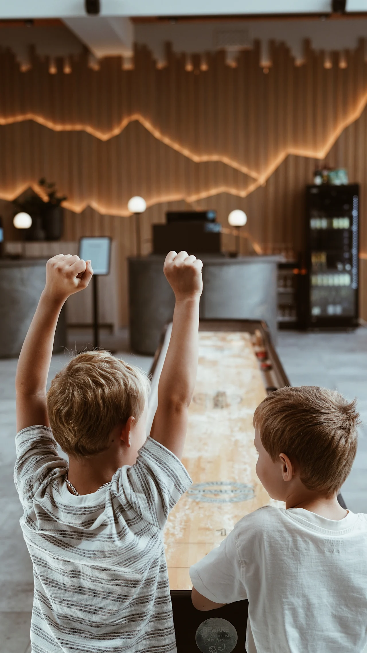 Barn spiller shuffleboard på Quality Hotel Arlanda XPO. Ett barn feirer et poeng. Synlig tekst: BEWARE The game consists of nutshells.