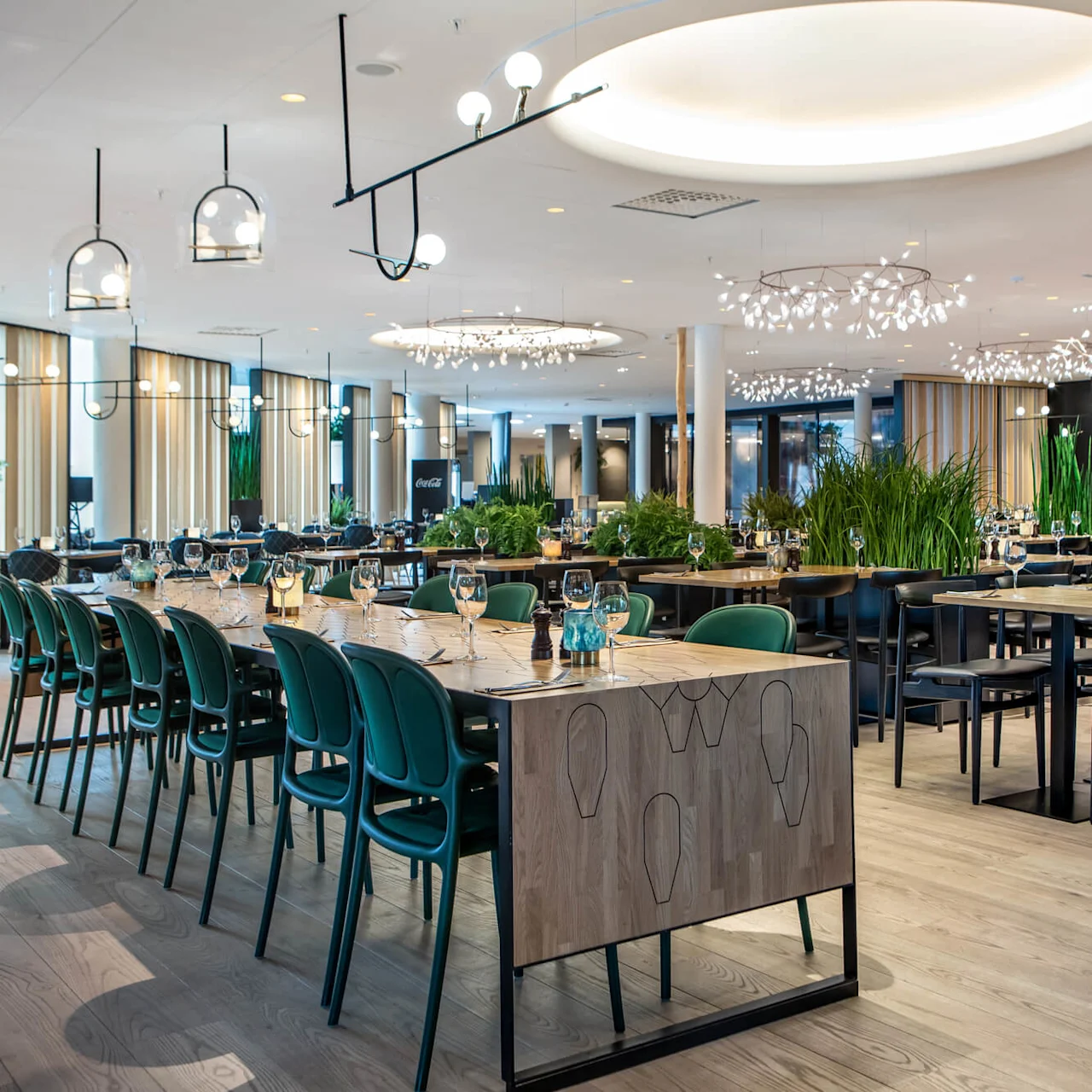 Luxurious dining area features elegant tables with green chairs, adorned with wine glasses, surrounded by lush plants, under modern chandeliers in a spacious, well-lit restaurant setting.