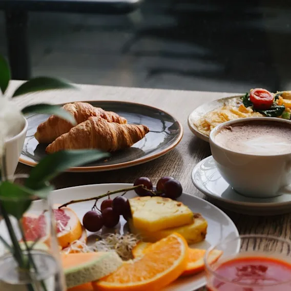 Breakfast spread with croissants, a plate of fresh sliced fruit, a cup of cappuccino, and juice on a wooden table.