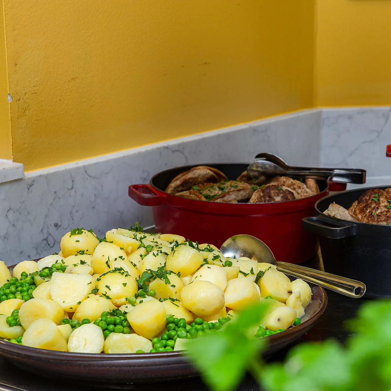 Kokade potatisar och gröna ärtor på en tallrik i förgrunden, köttbullar i kastruller bakom, placerade på en köksbänk mot en gul vägg.