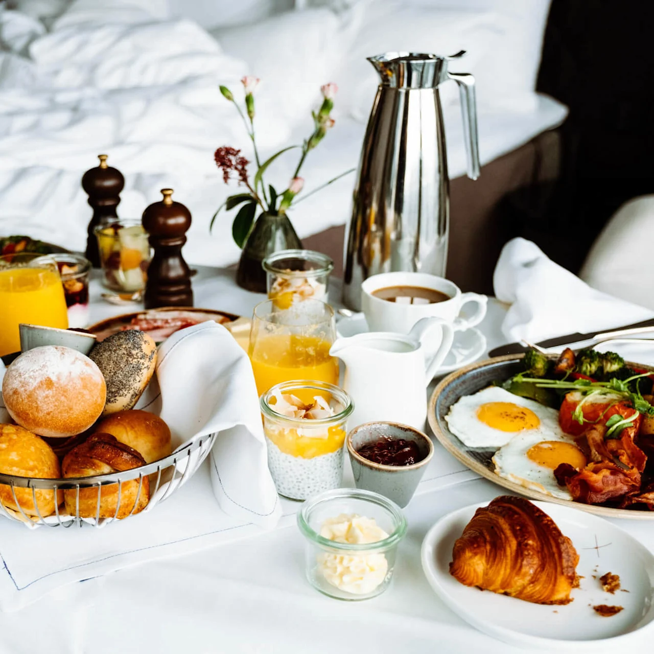 Frokostanretning på et bord inkluderer croissanter, rundstykker, egg, bacon, appelsinjuice og kaffe satt mot en uredd seng og små blomster i en vase.