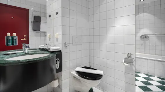 Vask med grøn bordplade ved siden af et toilet med foldet håndklæde, i et hvidflisebelagt badeværelse med håndklædeholder og synligt badekar. Flasker mærket "RITUALS".