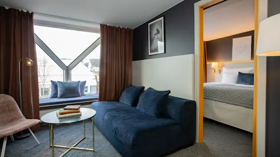 Ett mysigt hotellrum med en marinblå soffa vänd mot ett runt soffbord med böcker. Stora fönster med bruna gardiner belyser rummet. I det intilliggande rummet syns en välbäddad säng.