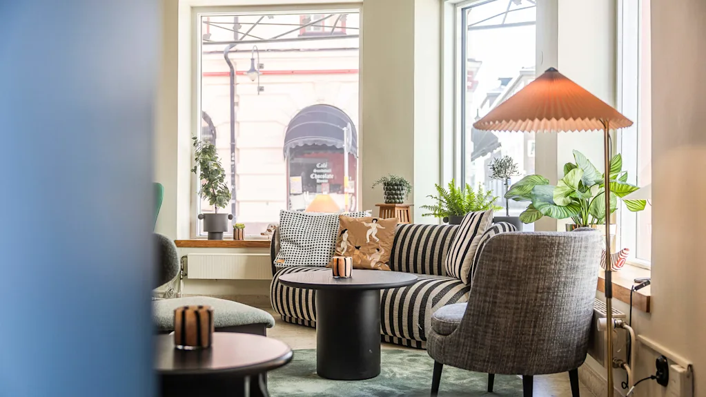 En mysig interiör med en randig soffa, växter, ett svart bord och en lampa, stort fönster med utsikt över gatan och ett café mittemot.