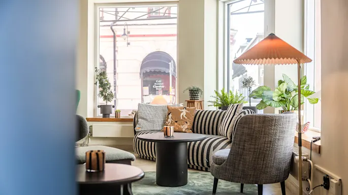 En koselig innvendig plass med en stripete sofa, planter, et svart bord og en lampe, med et stort vindu som ser ut over en gate og et kafé motsatt.