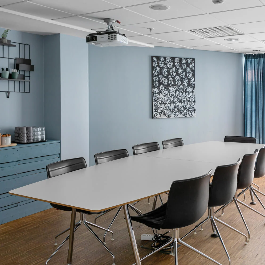 Ett konferensbord omges av svarta stolar i ett rum med blå gardiner och väggar. En tavla med abstrakta ansikten hänger på väggen.