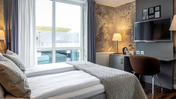 En prydligt bäddad säng med grå sängkläder och kuddar; bredvid, ett fönster. Ett skrivbord med en lampa, TV, vattenkokare och inramade dokument ligger i närheten. Allt i en modern hotellrumsmiljö.