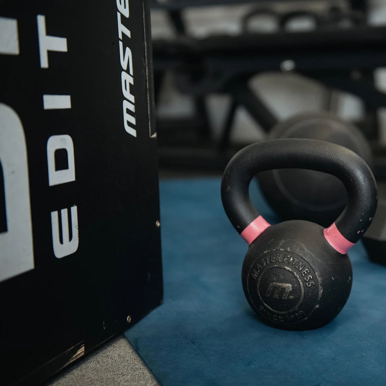 A black kettlebell labeled "Master Fitness" rests on a blue mat in a gym, accompanied by a dumbbell and a bench. Letters on a box partially read "EDIT" and "MASTER."