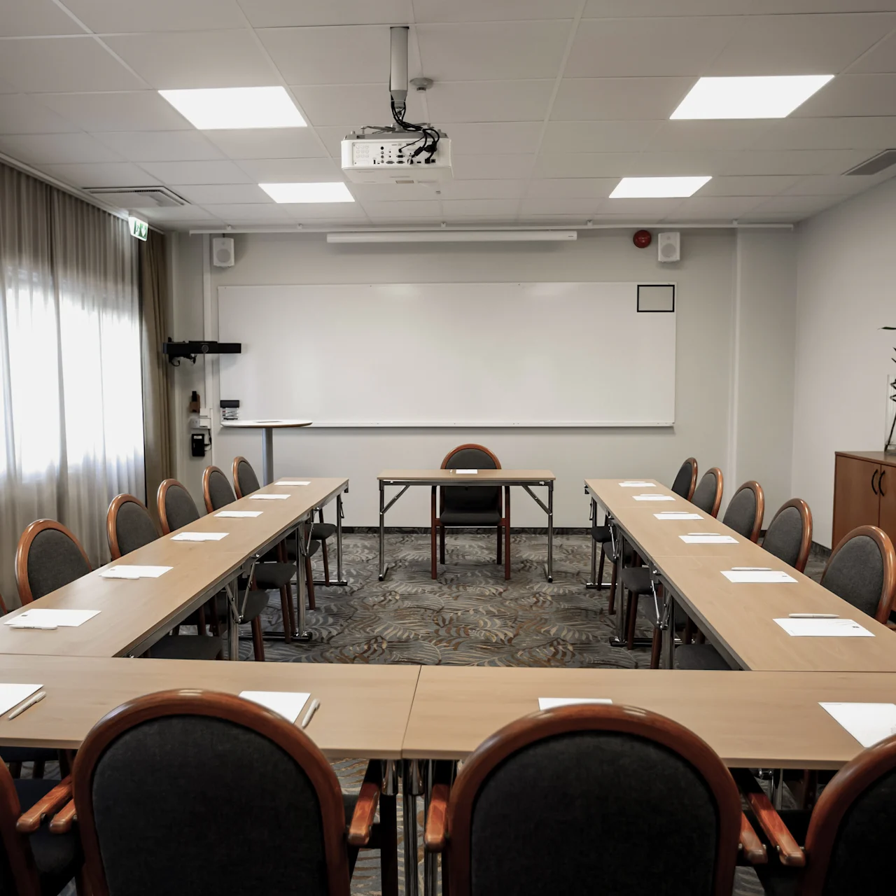 A U-shaped table setup with chairs surrounds a board, projector, and cabinet in a bright meeting room. Note pads and pens are placed on the tables.