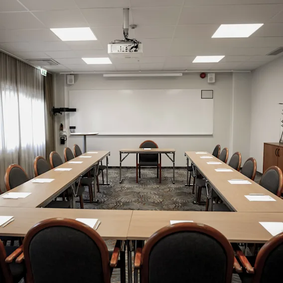 Ett U-format bord med stolar omger en tavla, projektor och skåp i ett ljust mötesrum. Anteckningsblock och pennor finns på borden.