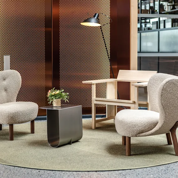 Komfortable kremfargede stoler og en trestol hviler på et sirkulært teppe med et lite metallbord imellom; en potteplante står på bordet, omgitt av en perforert vegg og en gulvlampe.