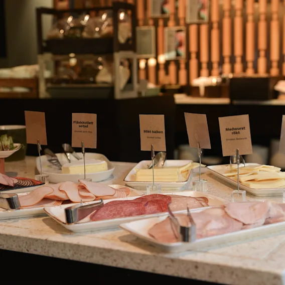 A selection of cold cuts and cheeses arranged on a countertop, each labeled with signs including "Vitvinskokt skinka," "Brieost," "Delikatess kalkon,&" and "Kallmarinerat fläskkött." Wooden decor is visible in the background.