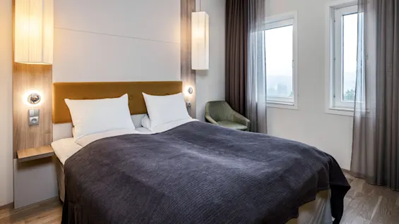 Seng med mørkegrått teppe; plassert i et moderne soverom. Vegger med to pendellamper, vinduer med grå gardiner, og en grønn lenestol i hjørnet.