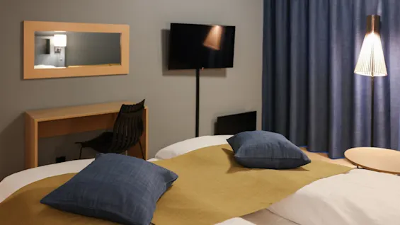 Två sängar med blå kuddar och en senapsgul filt finns i ett hotellrum. I närheten står en svart stol vid ett träskrivbord, med en väggmonterad TV och blå gardiner.