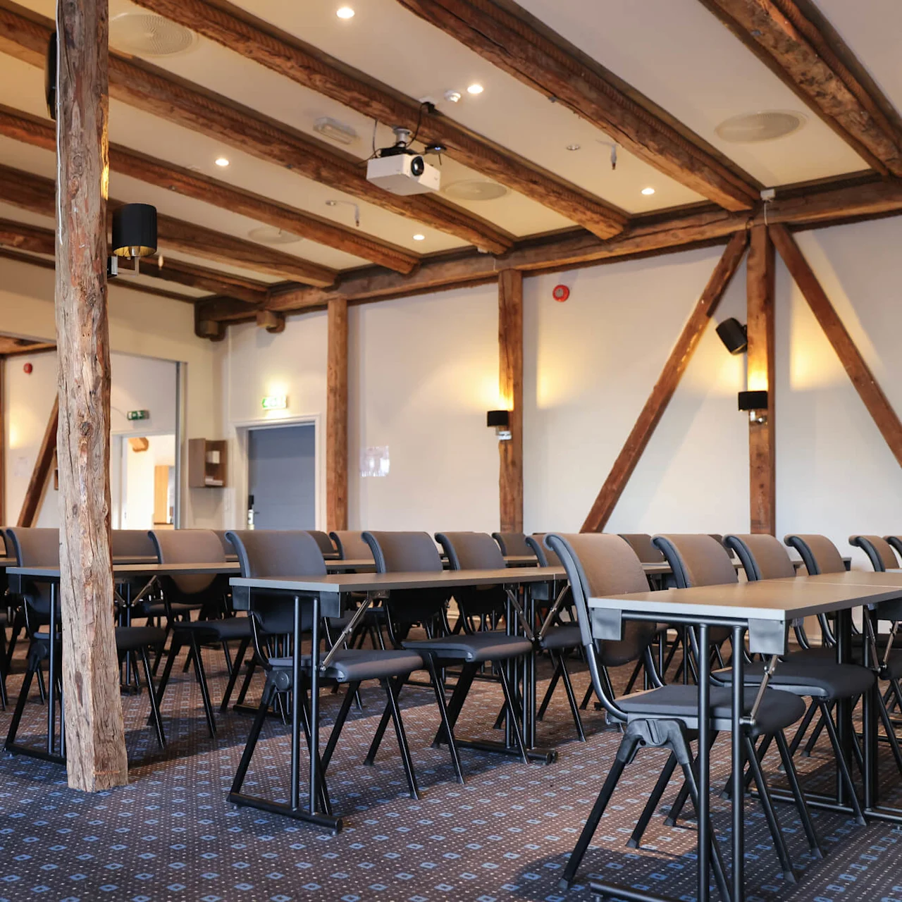 Rows of empty chairs and tables face a projection screen in a bright, wooden-beamed conference room. Walls are light-colored with black wall lamps.
