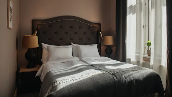 A neatly made bed with white pillows and a gray blanket is flanked by two lamps, situated in a warmly lit bedroom next to a large window with sheer curtains.