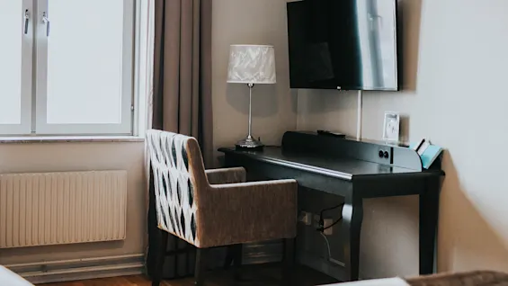 Et hotelværelses arbejdsområde med et skrivebord, en stol, en lampe og et tv ved siden af et vindue med gardiner.