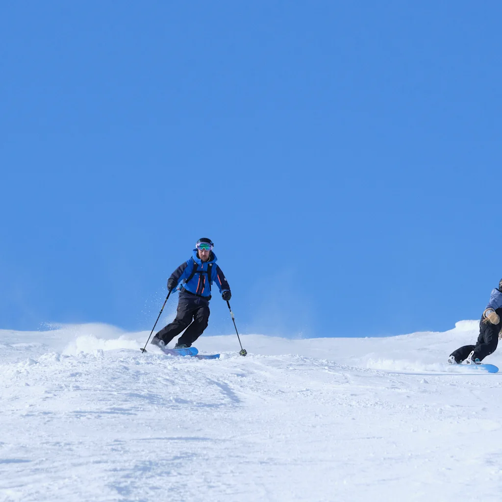To personer nyder vintersport på et sneklædt bjerg, den ene står på ski og den anden på snowboard under en klarblå himmel.