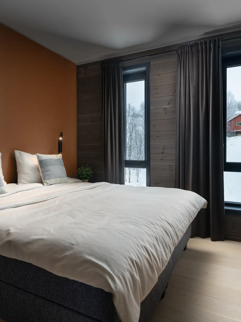 En prydligt bäddad säng med två kuddar står i ett mysigt sovrum med bruna väggar och stora fönster med utsikt över ett snötäckt landskap med ett rött hus.