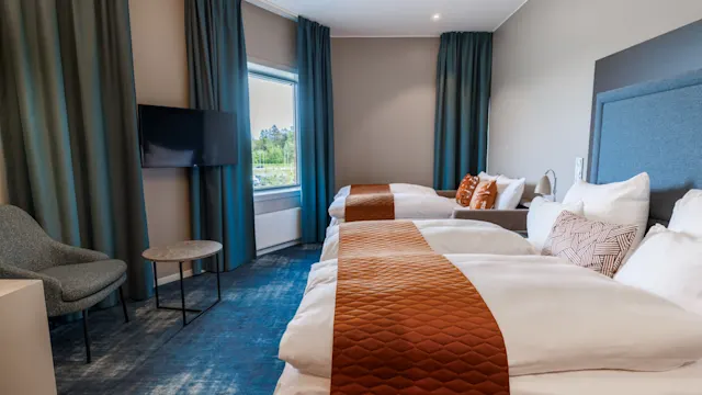 Kaksi siististi pedattua sänkyä, joissa on oranssit päiväpeitteet, sijaitsevat hotellihuoneessa. Televisio on seinällä, ja tuoli ja pieni pöytä ovat lähellä ikkunaa, jossa on verhot.