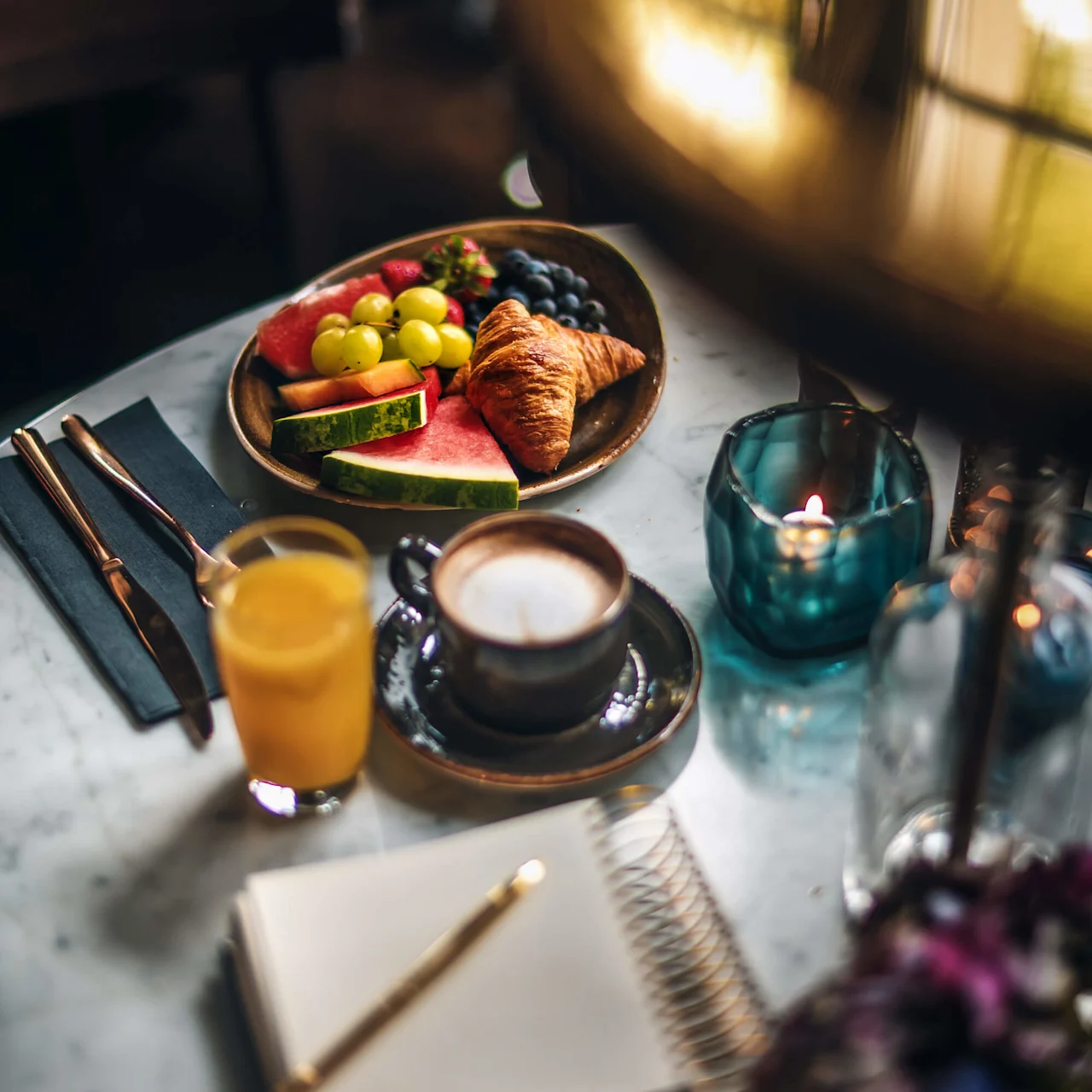 A plate rests on a marble table holding a croissant and various fruits. Nearby, there's a cup of coffee, a glass of orange juice, a notebook with a pen, and a lit candle.