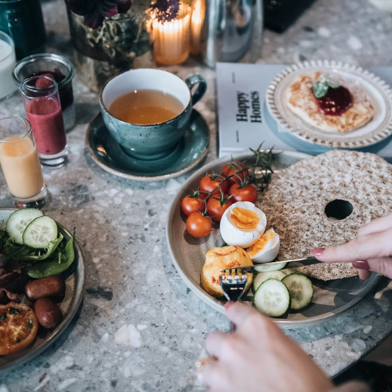 A person cuts soft-boiled eggs on a breakfast plate with crispbread, cherry tomatoes, and cucumber. Nearby are tea, vibrant smoothies, and a plate with pancake and jam on a magazine titled "Happy Homes."