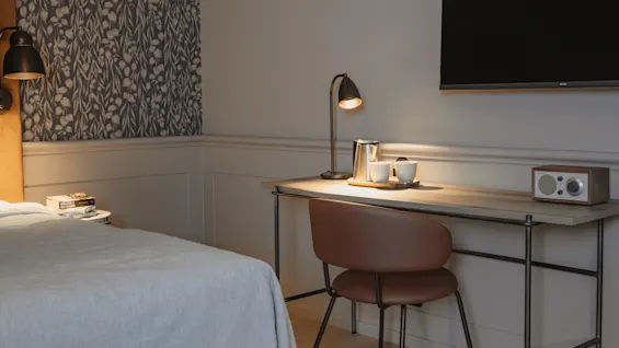 A modern room with a comfortable bed and a well-appointed desk area, featuring a lamp, kettle, and radio for guest use.