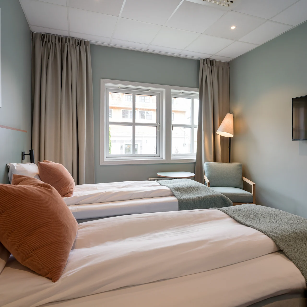 Two neatly made single beds with brown pillows, adjacent to large windows, a small table, and an armchair under a lit floor lamp; a television mounted on a pastel blue wall.
