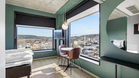 Et moderne hotellrom med et stort vindu som tilbyr panoramautsikt over en by, med en seng og et lite sitteområde.