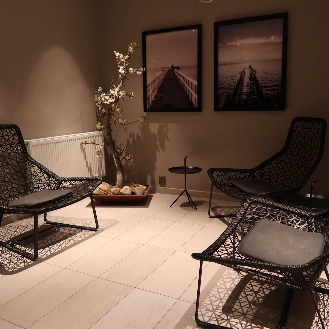 Four woven chairs encircle small tables in a dimly lit room. Two framed pictures and a decorative plant adorn the neutral walls, creating a calm, minimalist atmosphere.