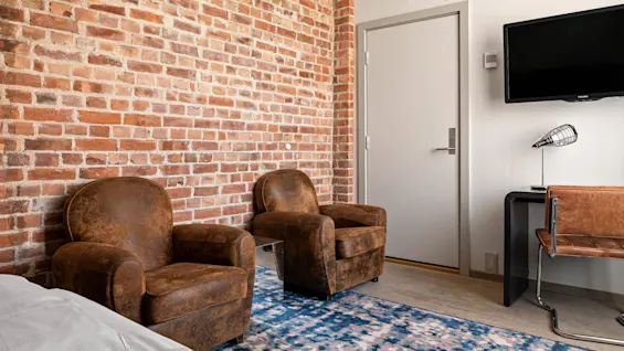 Two brown armchairs sit against a brick wall in a cozy room. A television and desk with a lamp are on the adjacent wall.