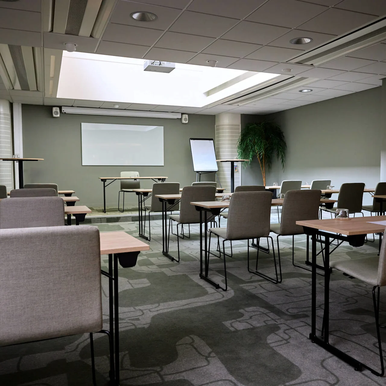 Meeting room with empty chairs and tables facing a whiteboard and flip chart; ceiling lights illuminate the space, which features a potted plant and patterned carpet.