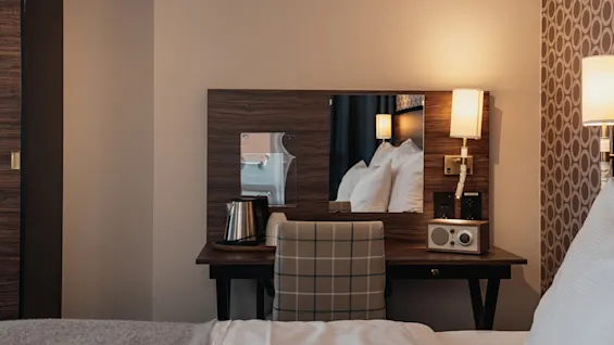 Viihtyisä hotellihuoneen nurkkaus, jossa on puinen peilipöytä, peili ja valikoima mukavuuksia, kuten vedenkeitin ja radio.