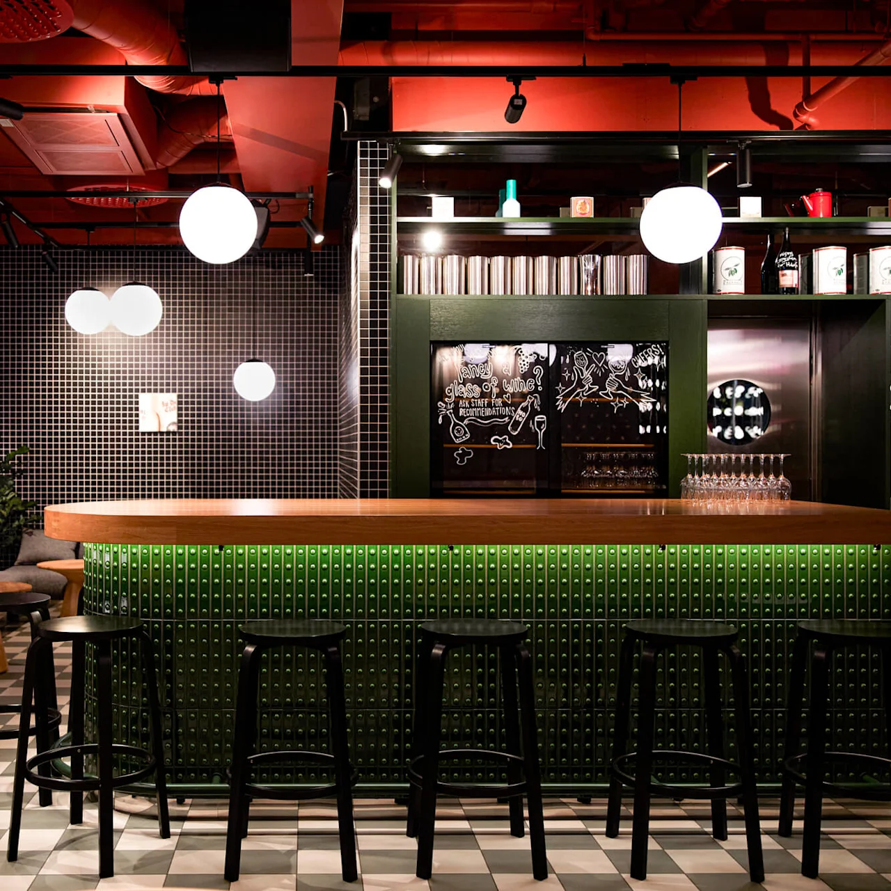 Bar med en trædisk omgivet af sorte stole, placeret i en moderne lounge med ternet gulv og mønstrede vægge. Cirkulære lysarmaturer hænger fra det røde loft.