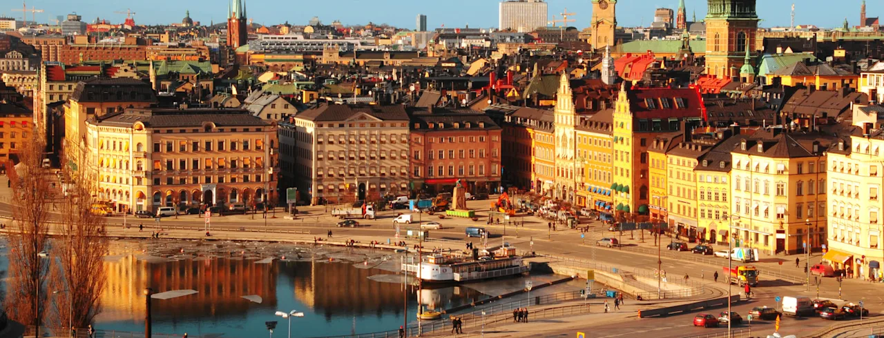 Colorful historical buildings line a bustling city square with people walking near a reflective waterway. Spires and towers rise against a clear blue sky, creating a picturesque urban scene.