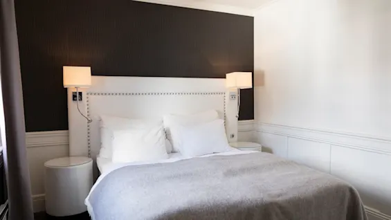 Et hyggeligt hotelværelse med en pænt redt seng, hvid sengegavl og sengelamper. Mørke og hvide vægge skaber en moderne stemning.
