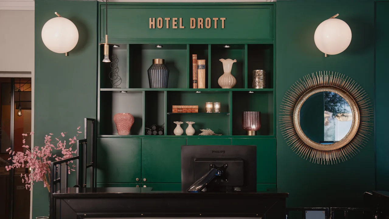 Reception med rosa blommor framför en grön hylla med vaser och böcker. "HOTEL DROTT" står skrivet ovanför. Runda speglar och lampor pryder väggarna.