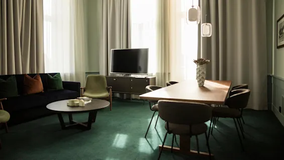 Ett stiligt vardagsrum med en mörk soffa och färgglada kuddar vänder mot en platt-TV på en skänk, omgiven av mjuka grå gardiner. Ett träbord med stolar kompletterar miljön.