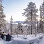Tre personer sidder på snedækket jord, omgivet af snedækkede træer, og ser ud over et fjernt snedækket landskab og bakker under en klar himmel.