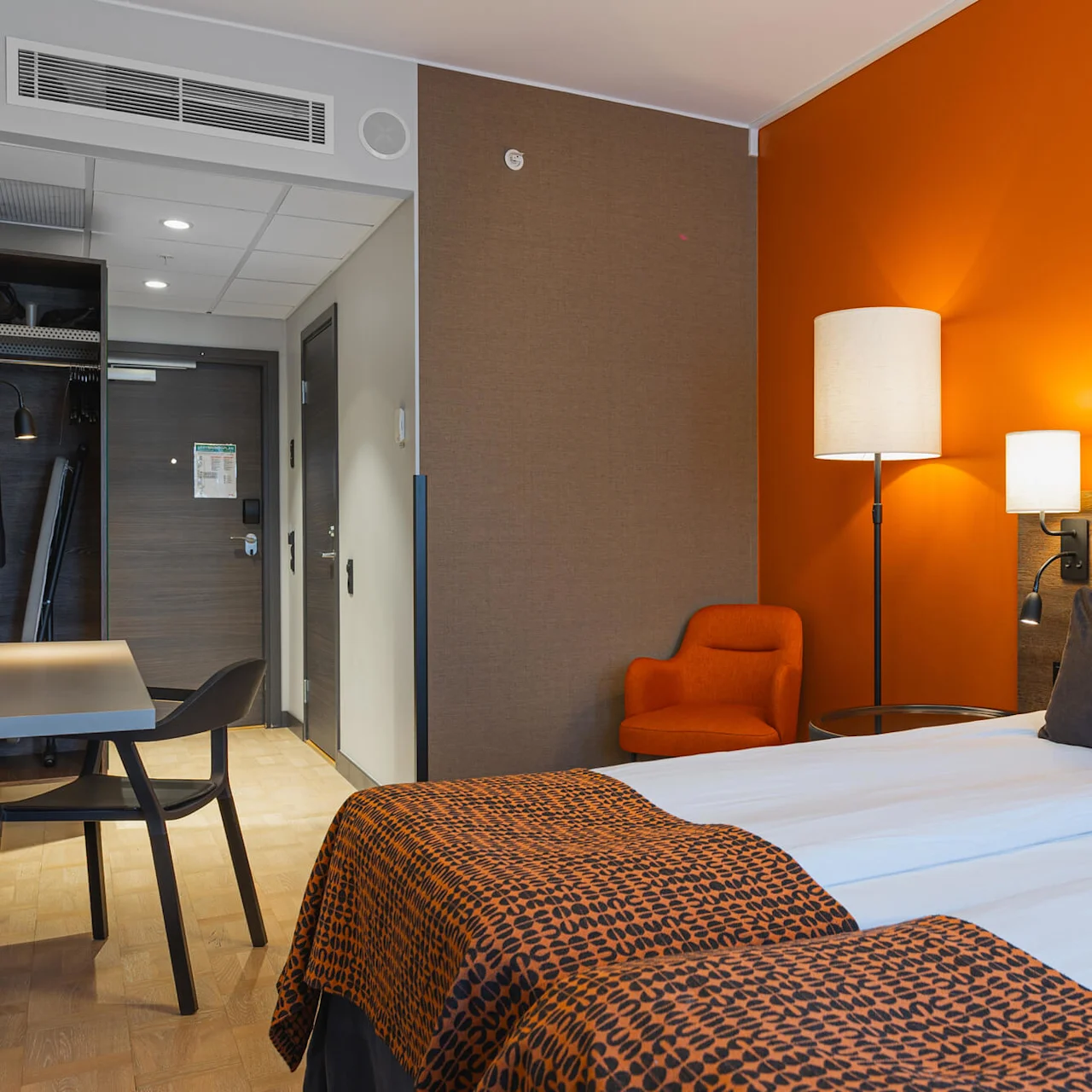 Ett hotellrum med en prydligt bäddad säng med orange och svart mönstrade överkast, en sänglampa, en orange accentvägg, en fåtölj och ett litet skrivbord.