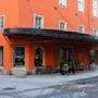En person trækker en kuffert forbi indgangen til en orange bygning med et sort udhæng, mærket "GRAND HOME HOTEL." Gaden er brolagt med sten.
