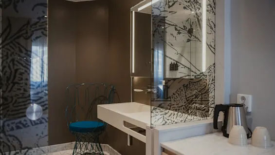 Ett elegant och funktionellt hotellbadrum med en mönstrad spegel, ett stilrent handfat och en livfull blå stol. En vattenkokare står redo.