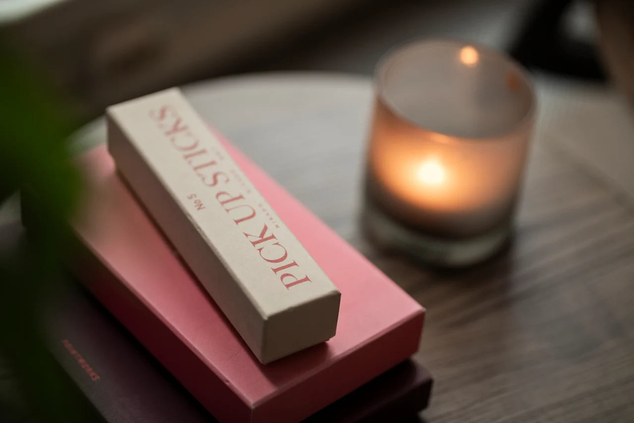 Rauhallinen ympäristö, jossa on laatikko PICK UP STICKS nro 5 ja pehmeästi hehkuva kynttilä, luoden rauhallisen tunnelman.