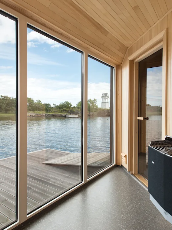 Innendørs sauna med store glassvinduer som vender mot en rolig innsjø med trær i det fjerne. Vegger og gulv i tre skaper en varm, fredfull atmosfære.