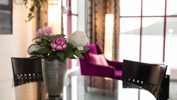Eloisa kukka-asetelma lasipöydällä valoisassa huoneessa, jossa on violetti nojatuoli ja luonnonkaunis ikkunanäkymä.