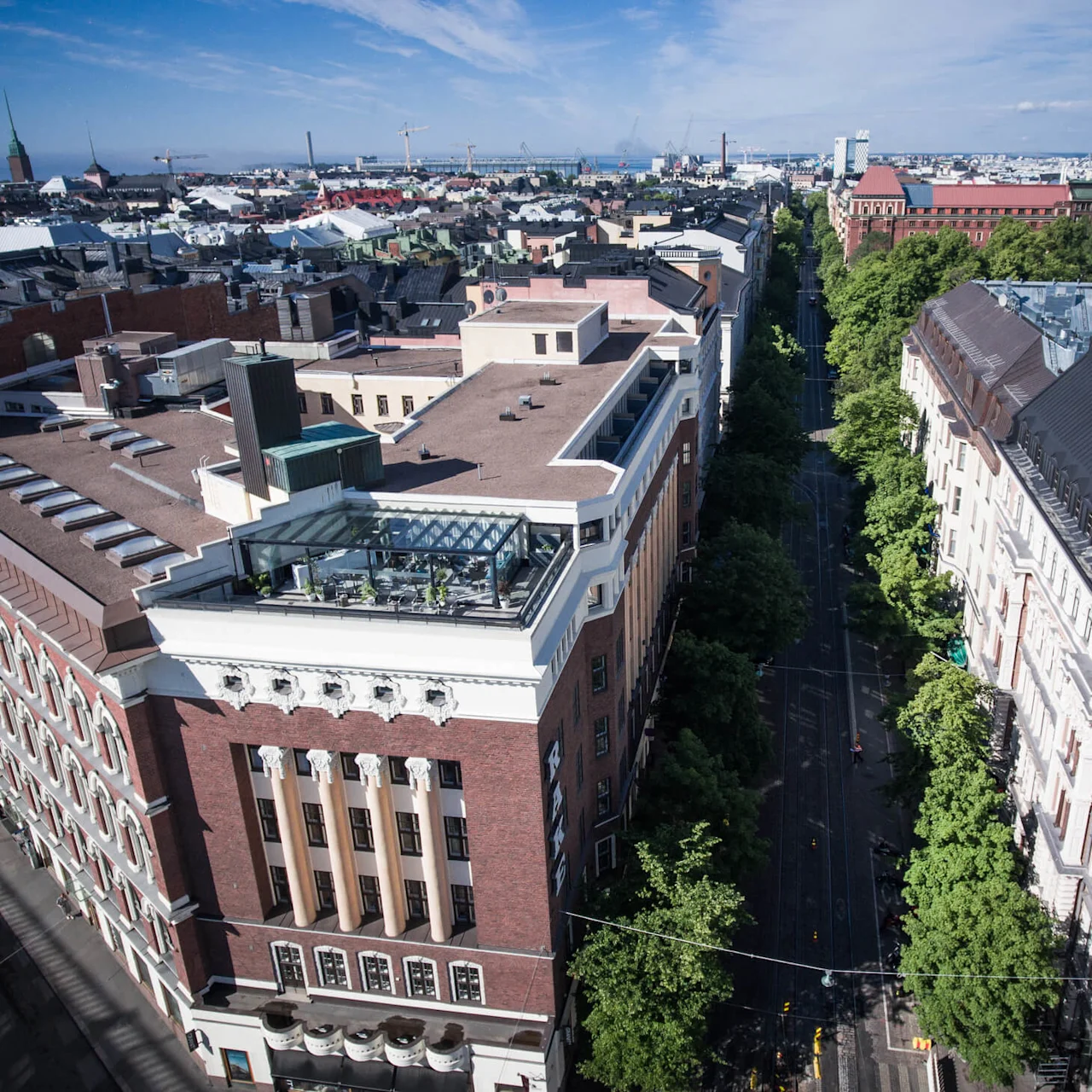 Ilmakuva kaupungista, jossa rakennukset reunustavat puiden reunustamaa katua, joka ulottuu kaukaiseen rantaviivaan. Katuterassit ja vanha arkkitehtuuri erottuvat kirkkaan sinisen taivaan alla.